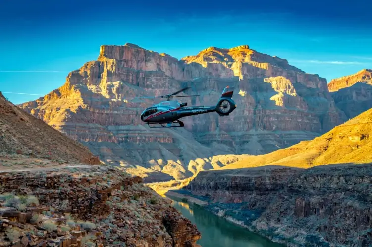 Helicopter flying over the Grand Canyon with the Colorado River below and sunlit canyon walls in the background.